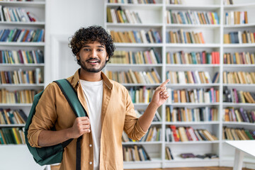 A happy young man with curly hair and a casual outfit is standing in a library. He is carrying a...