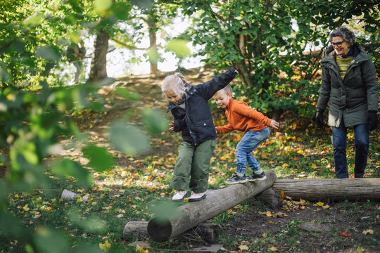 Female teacher looking at children balancing on wooden logs while playing at park