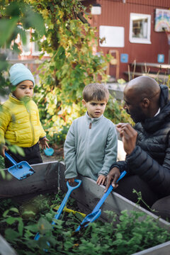 Male teacher assisting kids holding shovel while standing near plants at garden