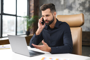 A businessman with a beard is sitting at a table in front of a laptop, talking on a cell phone, male office employee in smart casual wear is in a phone conversation