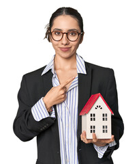 Caucasian businesswoman with house model, real estate concept on studio background smiling and pointing aside, showing something at blank space.