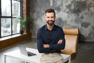 A confident businessman with a beard is sitting at a wooden desk with crossed arms, looking at the camera with a friendly smile. Male office employee in the workplace