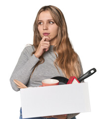 Blonde Caucasian woman moving with a box on studio backdrop looking sideways with doubtful and...