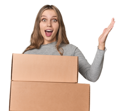 Blonde woman with cardboard boxes moving on studio backdrop receiving a pleasant surprise, excited and raising hands.