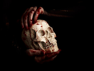 Bloody hands of a witch holding human skull over black background