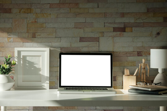 Laptop with blank screen, picture frame and stationery on wooden table against brick wall.
