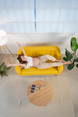 Top view of pregnant woman rests on yellow couch in a cozy living room happy with outstretched arms decorated with plants, surrounded by soft linens and bedding for comfort and relaxation