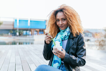 A woman with curly hair is sitting on a wooden bench and eating ice cream. She is wearing a black jacket and a blue scarf. The scene is casual and relaxed, on the outdoors of shopping mall. She's look