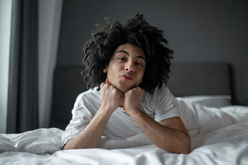 Fototapeta na wymiar Young curly-haired man lying in bed and looking relaxed and sleepy