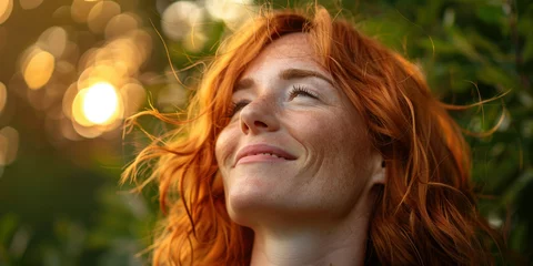 Poster Happy woman with red hair smiling and looking up at the sun in the background portrait in nature concept © SHOTPRIME STUDIO