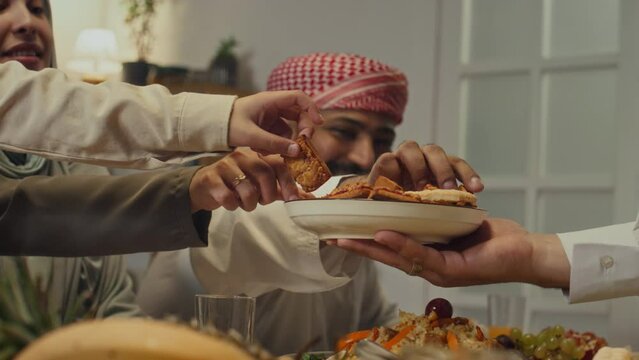 Side footage of unrecognizable hospitable host offering pastry or biscuits to male and female muslim family members during festive dinner on Eid al-Fitr