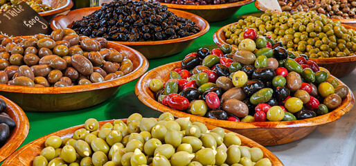 A close up of varied display of many types of fresh and dressed olives in wooden bowls, for sale at a supermarket or market.