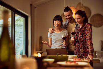 Happy friends using a laptop and talking to someone over the video call while drinking wine and having dinner together.