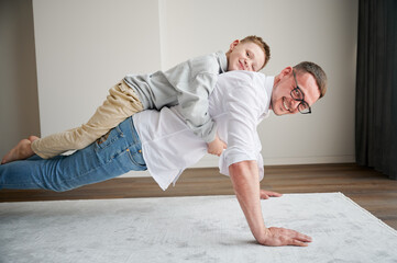 Energetic father and his little son training at home. Kid climbed dad's back while he doing plank. Young family enjoying activity games at home.