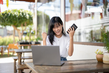 Fototapeta premium Happy Asian woman raising fist in victory while looking at her smartphone with a laptop in an outdoor cafe