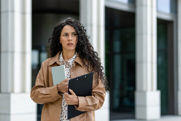 A professional woman stands outside a modern building, poised with a tablet and clipboard in hand....