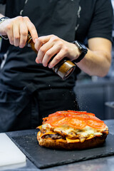 A chef meticulously seasons a freshly made sandwich with melted cheese and tomato slices on a black...