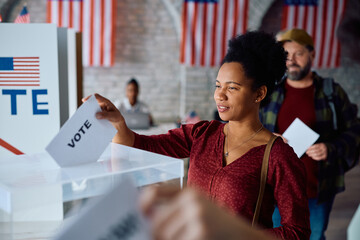 Black woman inserting her ballot into box while voting during US elections.