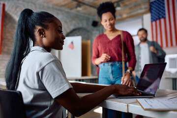 Black female volunteer working on laptop during voting at polling place.