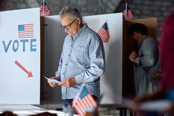 Senior man with a ballot at voting booth during US elections.