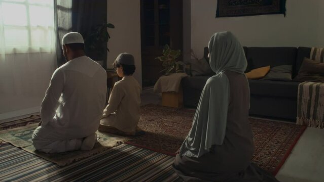 Rear footage of unidentified family of father, mother and son sitting on knees on floor and performing namaz at home