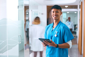 Portrait Of Smiling Male Doctor Or Nurse Wearing Scrubs In Hospital With Colleague In Background