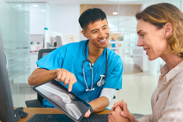 Male Doctor In Scrubs At Hospital Appointment With Female Patient Looking At Digital Tablet
