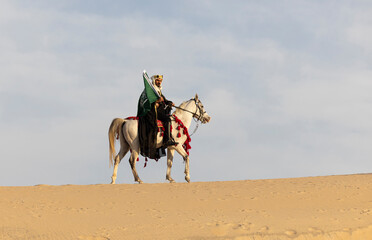 Rider, in traditional clothing, carrying a flag of Saudi Arabia on a white horse in a desert