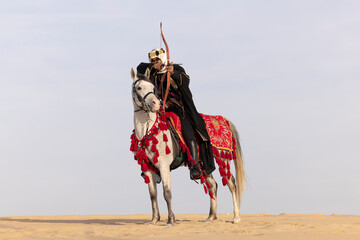 Saudi man in traditional clothing in the desert with a white horse, hunting with a bow and arrow
