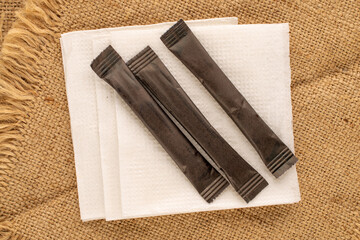 Several paper sugar sticks with paper white napkin on jute cloth, macro, top view.
