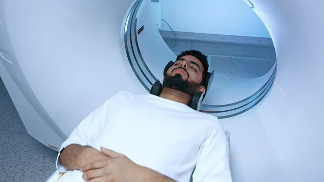 Caucasian handsome man lying on CT or MRI scanner during machine imaging body, lights up infrared rays and male patient passes through circle, crane shot from down to up, room interior.