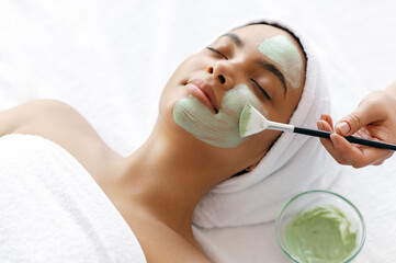 Top view of beauty procedure, therapist applying green face mask on the face of a beautiful brazilian or hispanic young woman. Facial treatments, wrinkle prevention, facial cleansing