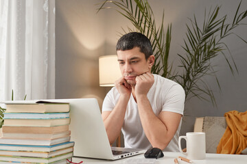 Bored sad Caucasian adult man wearing white T-shirt working at home with laptop looking at computer display with negative feeling has no mood and desire to work