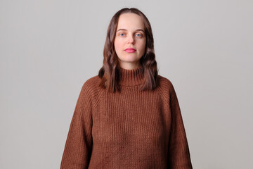 Sad serious Caucasian brown haired woman wearing brown jumper standing isolated over light gray background looking at camera with unhappy facial expression
