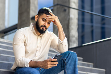 Photo of a sad young Muslim man sitting near a building on the stairs, holding his head and looking worriedly at the mobile phone screen