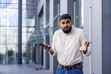 Portrait of a young Arab man standing on the street near an office building, holding a credit card and a mobile phone, looking worriedly at the camera and spreading his hands