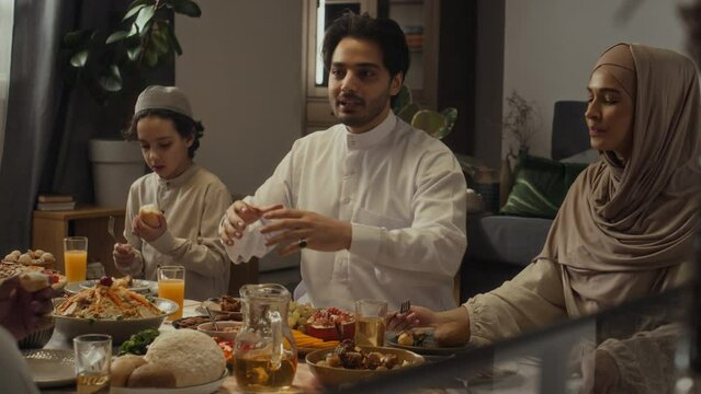 Side handheld footage of cheerful muslim man in thobe excitedly talking about some story while family members eating festive meal and listening to him at home