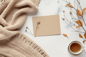 A white card with a flower on it sits on a table next to a cup of tea and a blanket mockup