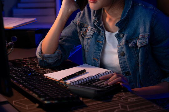 Cropped image of young Asian creative woman calling on phone to customer or coworker for marketing creator plan on pc with notebook and pen to write memo at neon modern office at night. Stratagem.