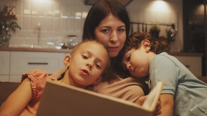 Mother enjoys reading storybook with her daughter and son, cuddling together on couch in cozy home setting, enjoying warm family moment