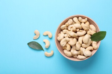 Many tasty cashew nuts and leaves on light blue background, top view. Space for text