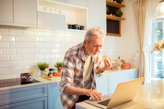 Senior man eating in the kitchen and using laptop