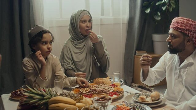 Handheld footage of muslim family members all taking nuts and dried fruits and enjoying then while speaking over festive table on Eid al-Fitr celebration at home