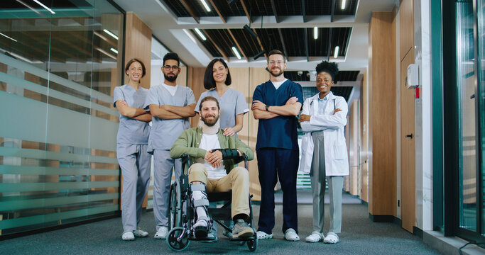 Portrait of successful diverse team of medical staff wearing uniform standing with crossed arms in corridor of modern hospital. Happy patient in wheelchair smiling. Teamwork concept.