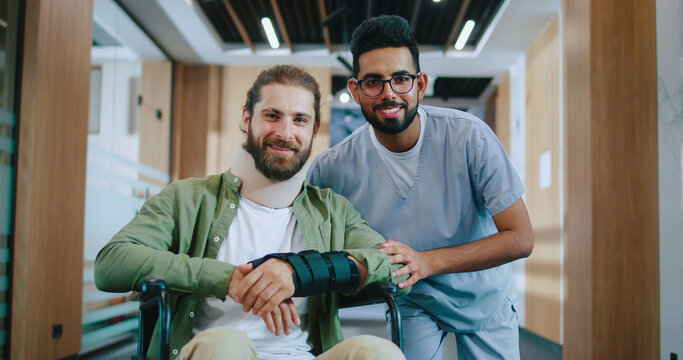 Portrait of happy Caucasian male patient injured in wheelchair recovering in modern hospital with professional Indian doctor in uniform. Handsome men looking at camera and smiling in hospital.