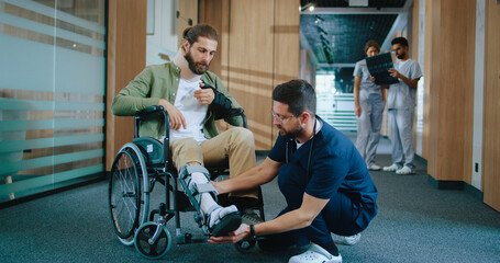 Young experienced Caucasian physiotherapist helping injured man in wheelchair with cast. Skillful qualified doctor in uniform consulting young patient in corridor of modern hospital.