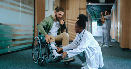 Caring successful African-American nurse helping injured patient in wheelchair. Young Caucasian man with cast talking with caring nurse in uniform. Modern hospital concept.