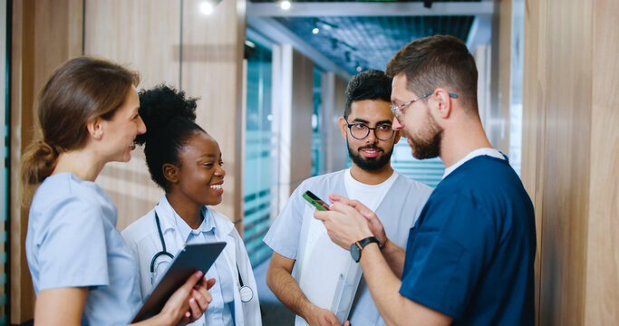 Side view portrait of diverse multi-age group of professional medical staff conversing in modern hospital corridor during break. Successful doctors and nurses using devices online.