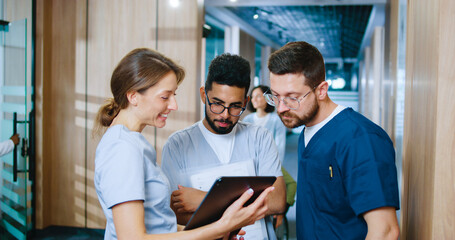 Side view portrait of multicultural group of young medical professionals chatting during break in contemporary hospital corridor. Nurses and doctors using gadgets online.