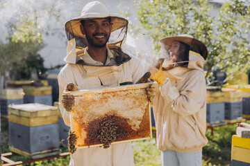 Happy Indian Beekeeper in a uniform standing in apiary and holding a honeybee frame on bees farm